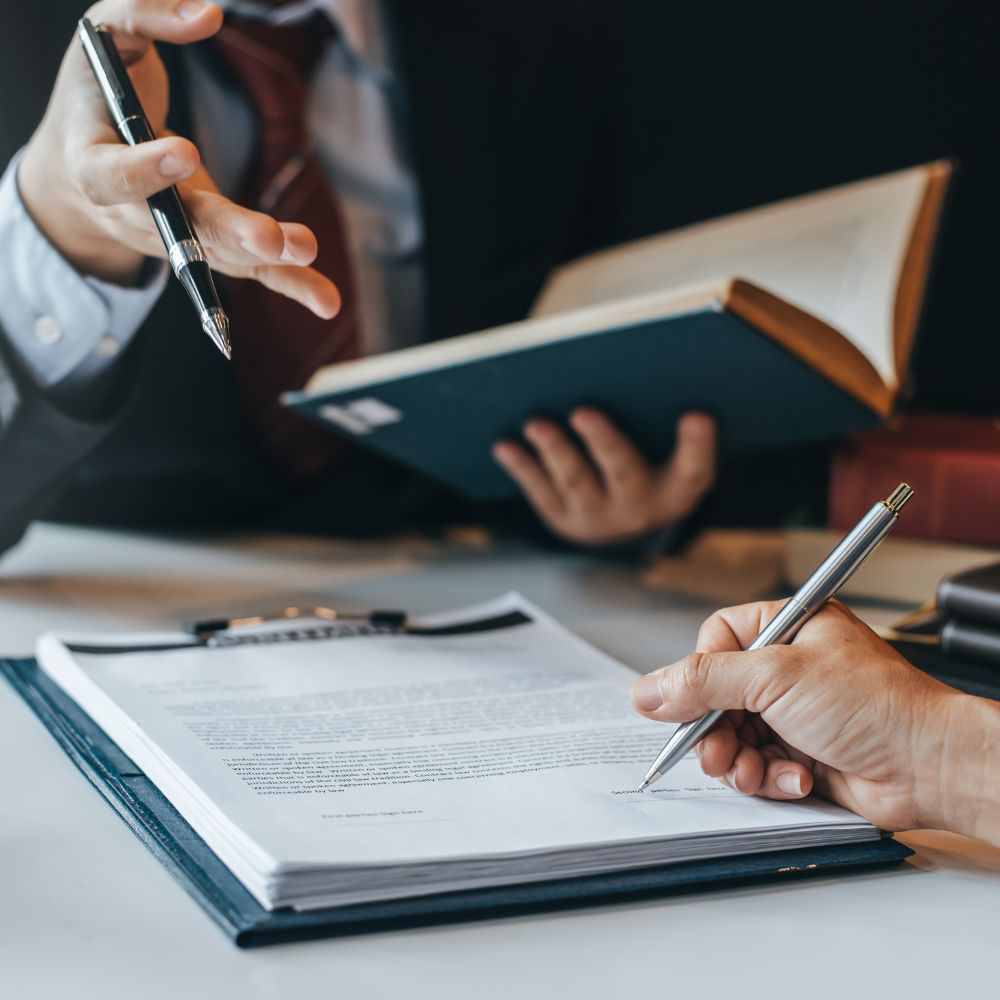 Close-up of hands signing a legal document while another person gestures during a meeting