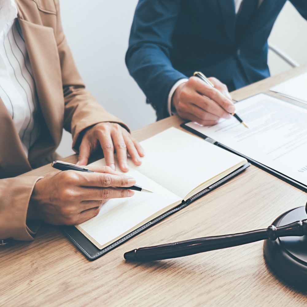 Two professionals in business attire writing notes and reviewing legal documents at a desk with a gavel in the foreground