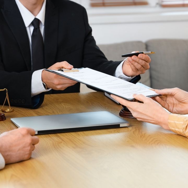 A lawyer holding a document and pen while another person reaches out to sign it at a table